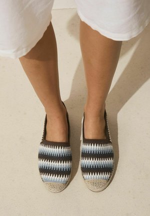 Legs wearing white shorts and slip-on shoes with blue, white, and brown zigzag patterns standing on a light-colored surface.