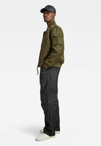 Veste bomber vert olive avec une fermeture éclair, des poches latérales et des poignets élastiques, portée avec un jean foncé et des baskets blanches, accompagnée d'une casquette noire.