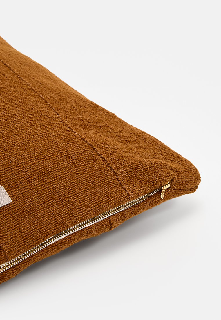 Kissen aus warmem braunem Textil mit sichtbarem Reißverschluss und einem kleinen Stoffetikett. Rechteckige Form, mit einem gewebten Muster.