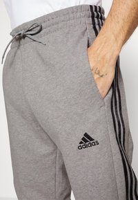 Grå sweatpants i mjukt tyg, med dragsko i midjan, sidofickor och svarta tre-strips detaljer. Adidas-logotyp i svart.