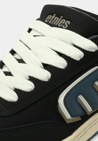 Μαύρο αθλητικό παπούτσι με λευκά κορδόνια, που διαθέτει μπλε λεπτομέρεια στο πλάι. Το υλικό φαίνεται να είναι συνδυασμός σουέντ και καουτσούκ.