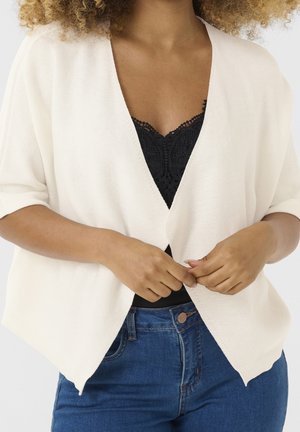 Femme portant un cardigan blanc ouvert sur un haut en dentelle noire et un jean bleu, mains jointes à la taille.