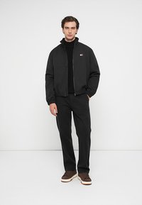 Sort bomberjakke med lynlås, ribbet manchetter, båret over en sort turtleneck. Sorte bukser og brune sko med hvide såler. Logo på jakken.