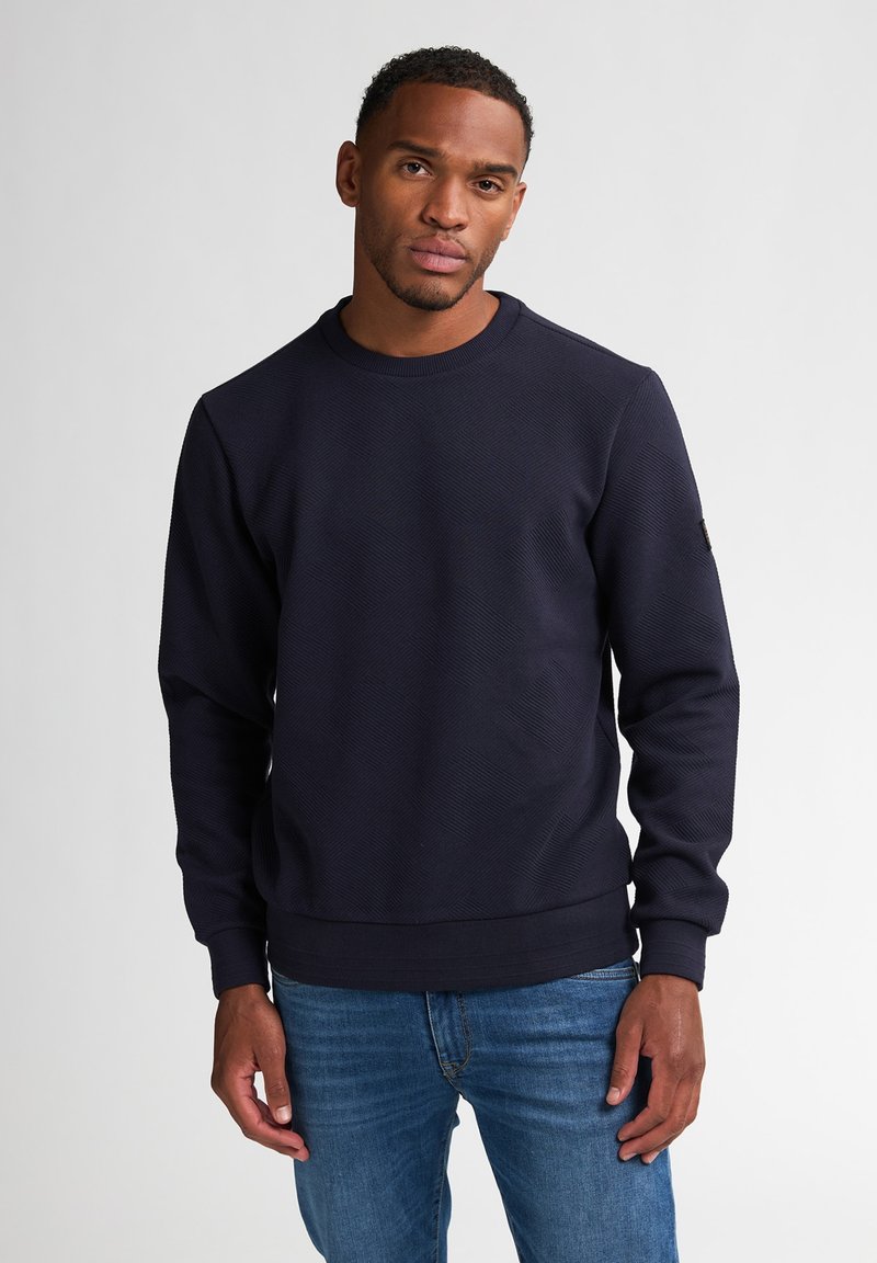 Felpa blu navy a girocollo con un motivo a diamante testurizzato, polsini e vita a coste, indossata con jeans blu.