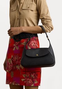 Mala de couro preta com uma textura suave e logótipo dourado, segurada por uma pessoa que veste uma camisa beige de botão e uma saia vermelha com padrão floral.