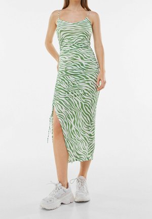 Femme portant une robe midi à rayures zébrées vertes et blanches avec des bretelles spaghetti et une fente latérale, associée à des baskets blanches épaisses.