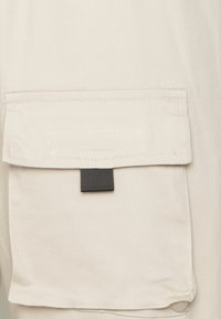 Beige stoffen cargopocket met een flap, voorzien van contrasterende donkergrijze banddetails en zichtbare stiksels. Gladde textuur met duurzame materialen.