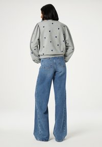 Žena s černými vlasy má na sobě šedou mikinu se vzorem malých tmavých srdíček a modré džíny s vysokým pasem a širokými nohavicemi, stojící na bílém pozadí.