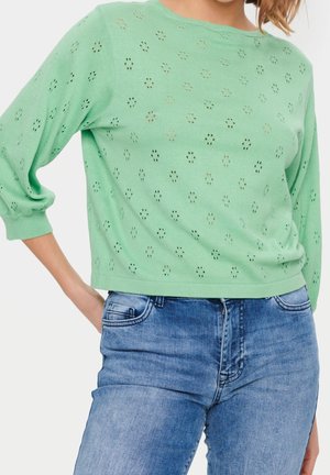 Femme portant un pull vert menthe avec de petites découpes florales et un jean en denim bleu, debout, une main sur la hanche.