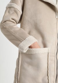 Abrigo de ante beige con detalles de piel de oveja en el cuello y los puños, que presenta un cierre de cremallera y bolsillos frontales con una textura suave.