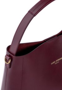 Burgunderfarbene Lederhandtasche mit glatter Textur, geschwungener Form und goldener Hardware-Akzent am Tragegurt; minimalistische Markenkennzeichnung sichtbar.