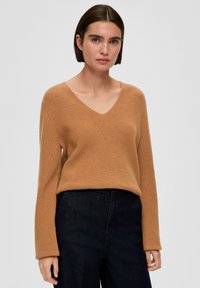 Sweter z dekoltem w serek, z długimi rękawami, w brązowym żebrowanym materiale, zestawiony z ciemnymi spodniami. Sweter ma luźny krój i teksturowaną tkaninę.