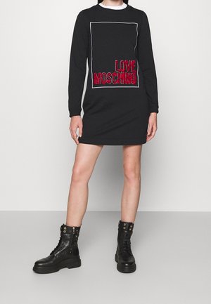 Schwarzes Langarmkleid mit rechteckigem weißen Umriss und rotem "LOVE MOSCHINO"-Text. Kombiniert mit schwarzen Schnür-Stiefeln im Kampfstil.