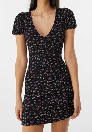Femme portant une courte robe noire avec un col en V et des manches bouffantes, présentant un petit motif floral rose et violet.