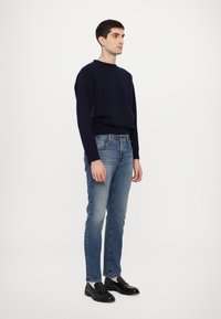 Un pull marine, un jean slim-fit bleu clair et des chaussures noires. Le pull possède un col rond et un ourlet côtelé ; le jean a un design classique à cinq poches.