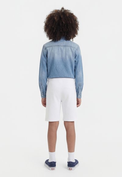 Personne aux cheveux bouclés de dos, portant une veste en jean bleu clair, un short blanc longueur genoux, des chaussettes blanches et des baskets bleu marine.