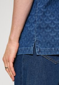 Chemise en denim avec une texture à motifs bleus, présentant un design à ourlet fendu. La chemise est associée à un pantalon en denim bleu foncé. Bague en argent visible à la main.