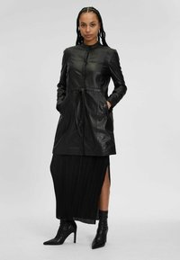 Cappotto in pelle nera con colletto, chiusura frontale con bottoni e vita con cintura. Presenta due tasche laterali e una texture liscia.