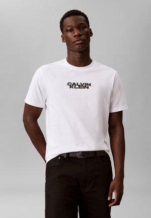 Homme portant un T-shirt blanc Calvin Klein et un pantalon noir, debout devant un fond gris uni.