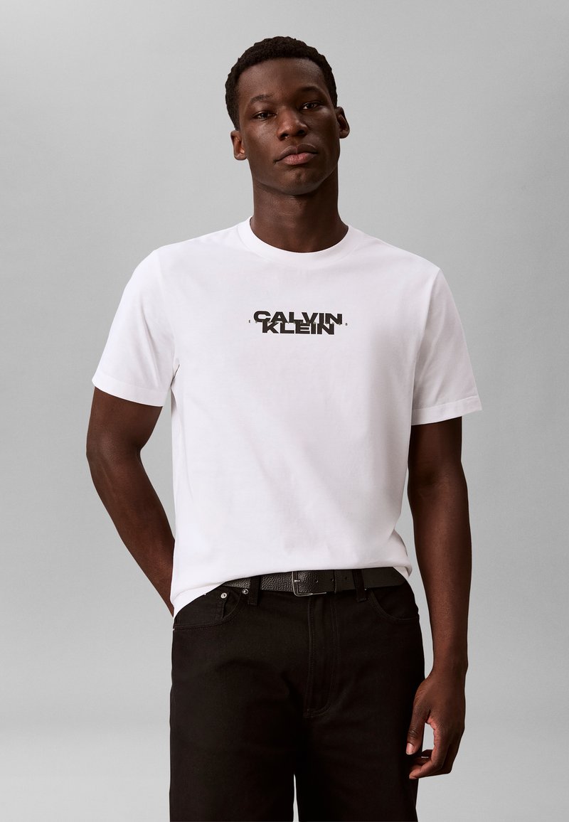 Άνδρας που φοράει λευκό μπλουζάκι Calvin Klein και μαύρο παντελόνι, στέκεται μπροστά σε απλό γκρι φόντο.