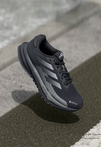 Juodas sportinis batas su tekstūrizuotais tinklelio viršutiniais, pilkų juostelių ir minkštu padu, turintis GORE-TEX etiketę vandens atsparumui.