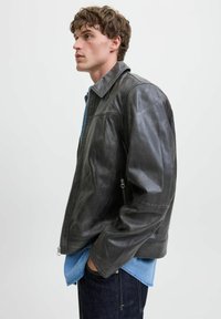 Chaqueta de cuero gris con una textura suave, que presenta un cuello alto, cierre frontal y bolsillos laterales. Lleva puesta una camisa de mezclilla azul.