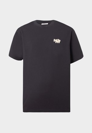 Ung mand står iført en sort T-shirt med korte ærmer, lysegrå bukser med vide ben og sorte sko, hænderne i lommerne.