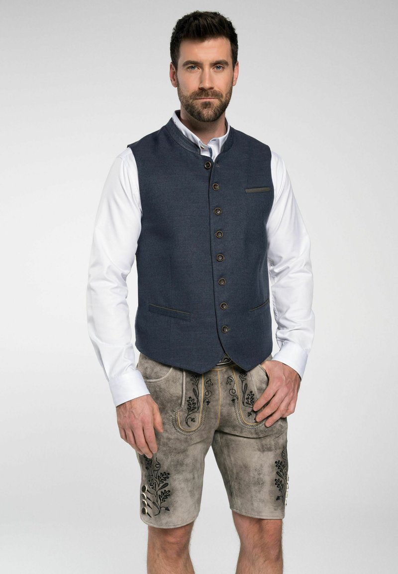 Marineblaues Weste mit hohem Kragen, Knopfverschluss und Fronttaschen, kombiniert mit grauen kurzen Lederhosen mit bestickten Akzenten.