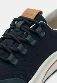 Αθλητικό παπούτσι από ναυτικό ύφασμα με υφή στην επιφάνεια, λαστιχένιες λεπτομέρειες και ταμπελάκι από δέρμα σε ταμπά χρώμα. Διαθέτει μοναδικό σύστημα δέσιμου με κουμπί και λευκή σόλα.