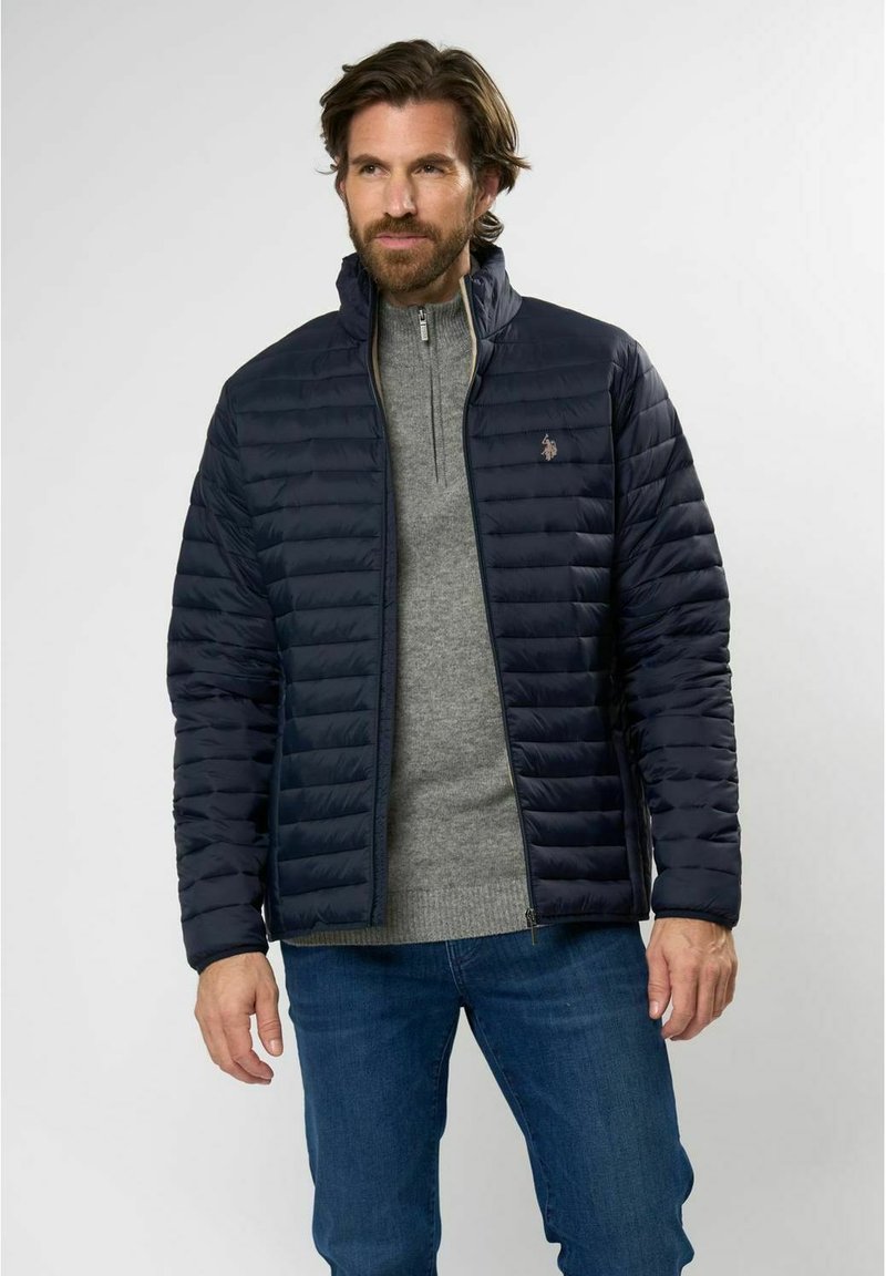 Marineblå puffjakke med glidelås foran, horisontal quilting og høy krage, båret over en grå genser, parret med blå jeans.