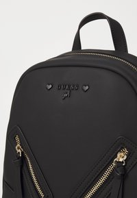 Sac à dos synthétique noir avec fermetures éclair dorées, logo "GUESS girl" en relief et accents en forme de cœur ; design géométrique avec des côtés texturés.