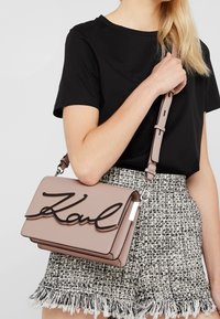 Różowa skórzana torebka z klapą, na której widnieje wyraźne czarne logo "Karl". Torebka ma odpinany pasek i gładką fakturę.