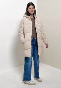 Manteau duffle beige avec capuche, design matelassé, deux poches avant, porté sur un pull marron et un jean large bleu, associé à des baskets blanches.