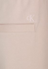 Tejido de color rosa claro con una textura suave, que presenta un discreto logo de CK bordado en el mismo color y un pequeño detalle de bolsillo.