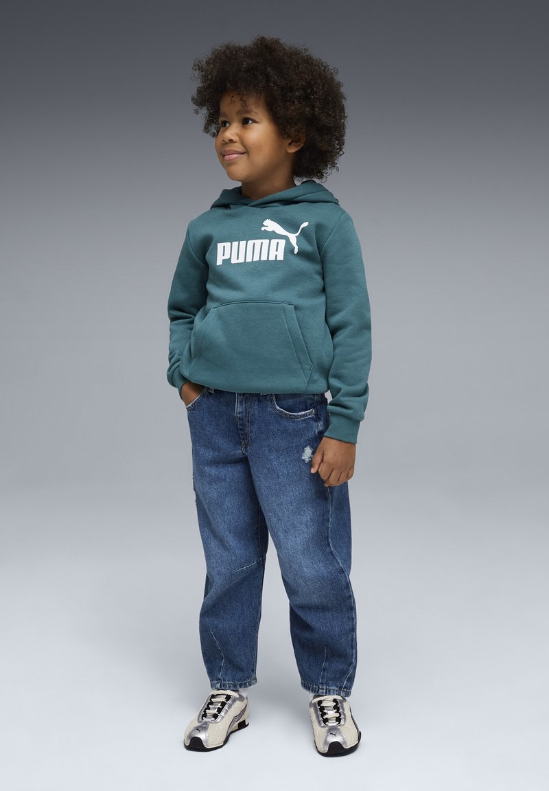 Sweat à capuche vert avec logo "PUMA" blanc, poche avant, assorti à un jean bleu déchiré et des baskets métalliques. Le modèle pose devant un fond gris.