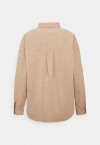 Chemise beige à manches longues avec un empiècement au dos, un pli creux central, des poignets boutonnés et un col classique sur un fond blanc.