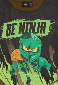 Postać ninja z Lego w zielonej zbroi z intensywnymi oczami i pomarańczowym naramiennikiem, z odważnym napisem "BĄDŹ NINJĄ" na zielonym tle z czarnym tłem.