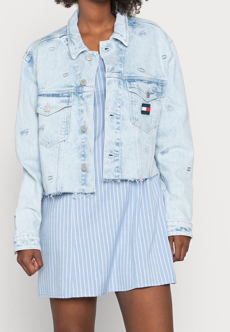 Ljusblå denimjacka i cropped-stil med slitna detaljer, silverknappar, två bröstfickor och en liten logopatch. Bärs över en randig klänning.