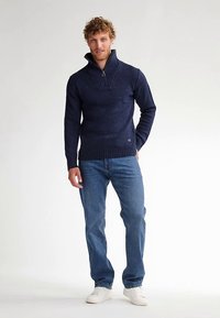 Laivastonsininen neule, jossa korkea kaulus, teksturoitu neulos ja vetoketju. Yhdistetty sinisiin farkkuihin ja valkoisiin lenkkareihin, miehen yllä, jolla on kiharat hiukset.