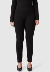 Pantaloni neri elasticizzati con una texture liscia, dotati di cuciture laterali, gambe affusolate e zip alla caviglia, abbinati a tacchi a punta neri.
