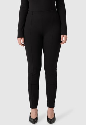 Zwarte stretchbroeken met een gladde textuur, voorzien van zijnaad, taps toelopende benen en ritssluitingen bij de enkels, gecombineerd met zwarte pumps met een spitse neus.