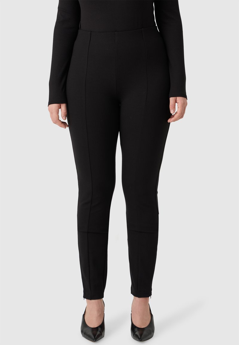 Pantaloni neri elasticizzati con una texture liscia, dotati di cuciture laterali, gambe affusolate e zip alla caviglia, abbinati a tacchi a punta neri.