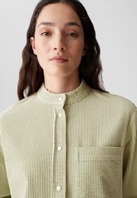 Camicia in corda verde chiaro con colletto alla coreana, strisce testurizzate, tasca frontale e bottoni a pressione lungo la patta.