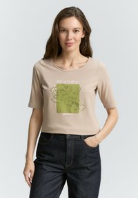 Beige T-Shirt mit kurzen Ärmeln, grünem Blumenmotiv und dem Text "BLOOMING". Weicher Stoff, runder Ausschnitt und lockere Passform.