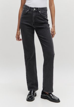 Schwarz-hochbundige Jeans mit geradem Beinschnitt, ausgestattet mit fünf Taschen, einem Knopfverschluss und klassischer Denim-Textur. Glänzende schwarze Slipper runden den Look ab.