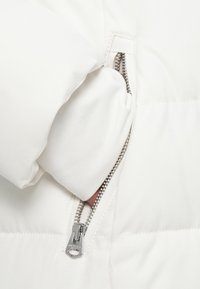 Χέρι σε λευκή πούφερ τζάκετ, μέσα στη τσέπη, με μερικώς ανοιχτό ασημί φερμουάρ σε λευκό καπιτονέ ύφασμα.