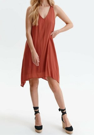 Frau trägt ein ärmelloses, rostfarbenes Kleid mit V-Ausschnitt und schwarze Keilsandalen mit Knöchelriemen.