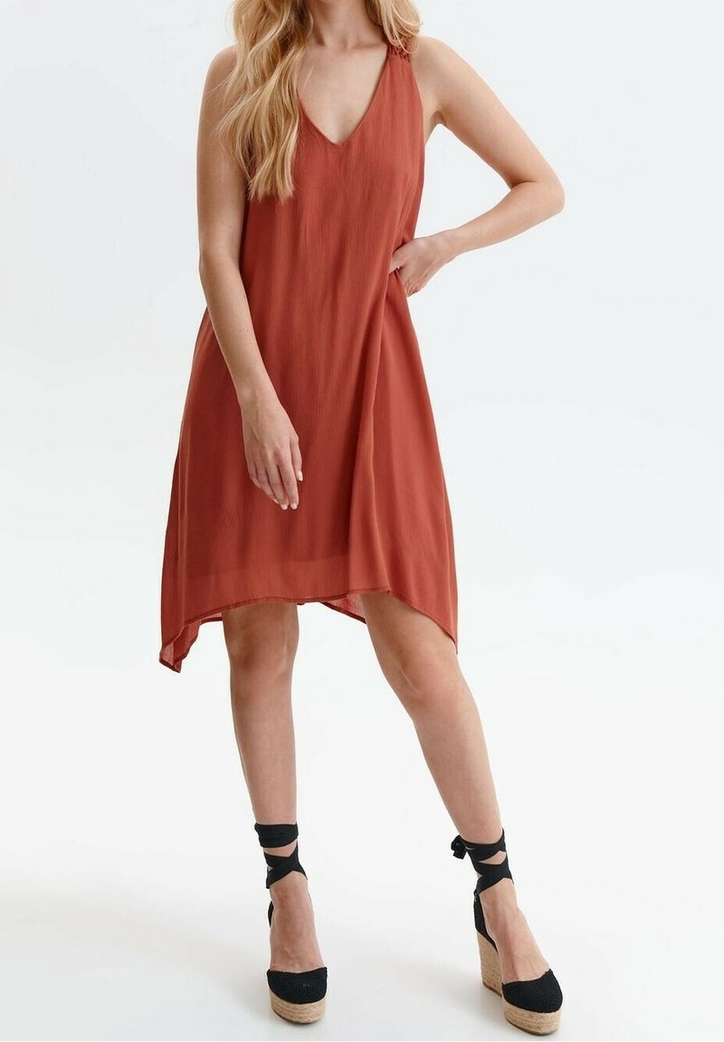 Femme portant une robe sans manches couleur rouille avec un décolleté en V et des sandales compensées noires avec des brides autour de la cheville.