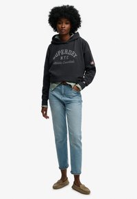 Ciemnoszara krótka bluza z kapturem z logo "SUPERDRY", nałożona na jasnozieloną koszulę, w zestawie z jasnoniebieskimi dżinsami i beżowymi butami wsuwanymi.