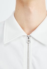 Polo blanc à fermeture éclair, avec un col pointu et un tissu texturé. Le tirette de la fermeture éclair affiche la marque « WEEKDAY ».